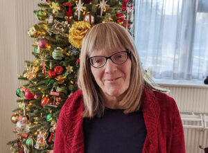 Woman with shoulder length blonde hair, wearing glasses, a red cardigan and black top in front of Christmas tree