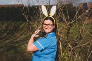Woman in blue nurse's uniform wearing white bunny ears, holding a black rabbit. In top left corner, an illustration of colourful flowers