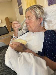 Blonde woman eating bowl of custard in hospital bed