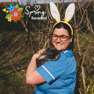 Woman in blue nurse's uniform wearing white bunny ears, holding a black rabbit. In top left corner, an illustration of colourful flowers with white text "Spring forward"