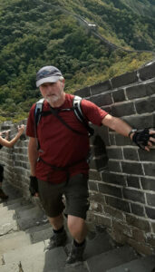 Man wearing grey hat and red T-shirt and black gloves on Great Wall of China, holding the wall, with mountains in background