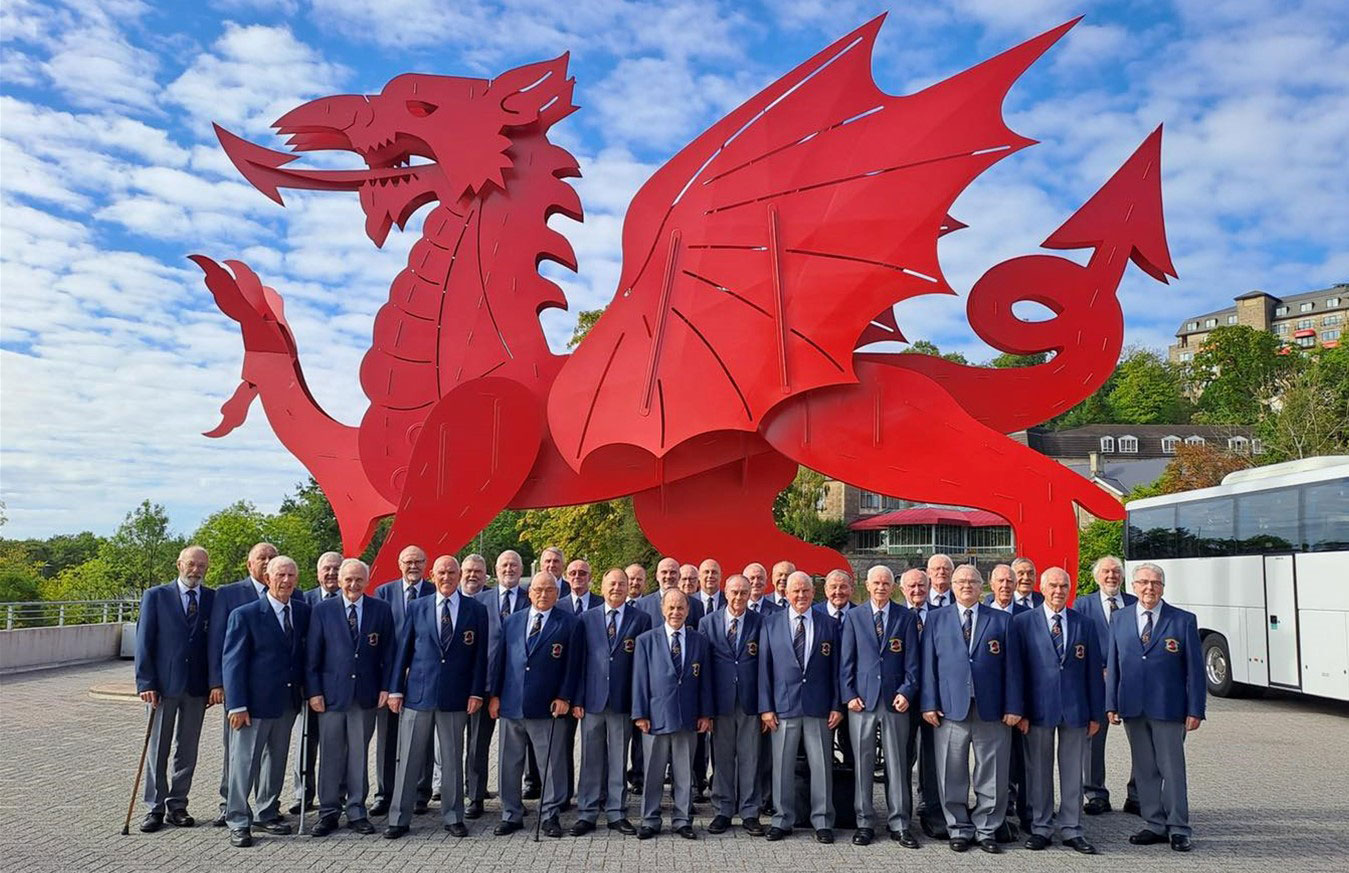 Large group of men wearing navy blue jackets and grey trousers in front of large red dragon statue, representing Wales