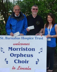 Two men and a woman with large white sign with text "St Barnabas Hospice Trust welcomes Morriston Orpheus Choir" in outdoor setting