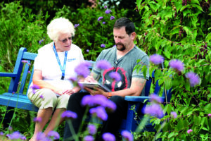 Am elderly woman wearing a white St Barnabas Hospice T-shirt and blue lanyard sitting on a blue bench with a dark haired man. They are surrounded by plants and trees, and are looking down at a notepad the man is holding.