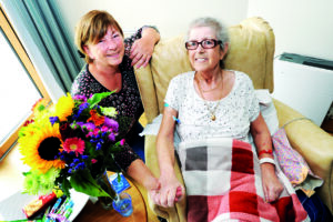Two women, one with short brown hair and the other with spiky grey hair, holding hands. The woman on the right is sitting in a beige chair with a red and grey checked blanket on her legs. Next to them is a vase with colourful flowers.