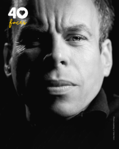 A man, Warwick Davis, who is Patron of St Barnabas Hospice, photographed in black and white, against a black backdrop