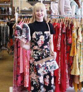 Blonde woman wearing floral jumpsuit holding shoes and bag, with red and yellow clothing on rack behind her