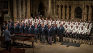 Group of men in white jackets sitting on chairs, group of men in black suit jackets standing by piano and singing at Lincoln Cathedral
