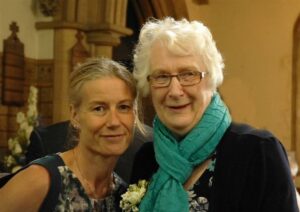 Two women smiling for camera. Louise Southgate on the left, a blonde woman wearing floral top and on the right grey-haired woman, Pam Burton, wearing green scarf and black cardigan