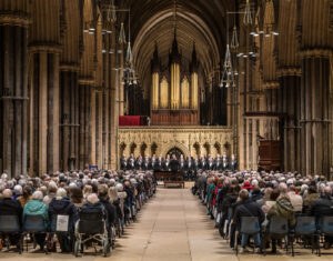 Wide angle view inside of Lincoln Cathedral with large organ against back wall and gothic arches. Audience sitting on chairs watching choir concert