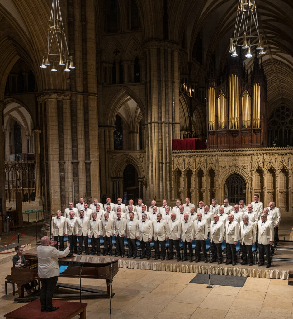 Wide shot of interior of Lincoln Cathedral with organ, and large group of men wearing white suit jackets, piano and conductor