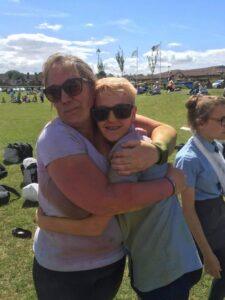 Blonde woman wearing white T-shirt and black trousers holding young girl with short hair and sunglasses in outdoors setting, hugging.