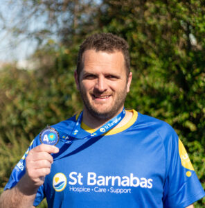 A man wearing a St Barnabas running top, holding a medal, stood in front of green foliage.