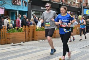Woman running Manchester Marathon, wearing blue St Barnabas running top, next to man wearing grey T-short