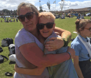 Blonde woman wearing white T-shirt and black trousers holding young girl with short hair and sunglasses in outdoors setting, hugging.