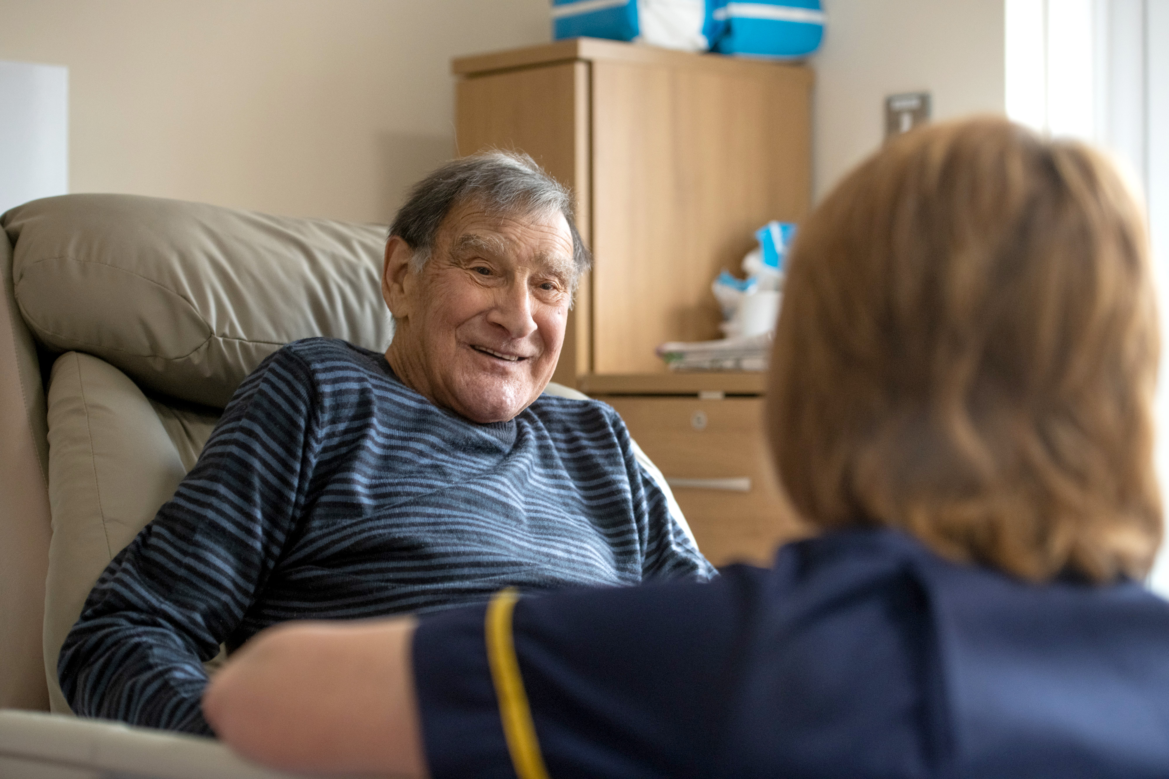 A man, sitting in a patient bed, smiling, whilst talking to a nurse.