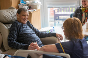 A man, sitting in a patient bed, smiling, whilst talking to a nurse.