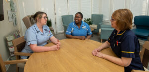 Three women, in nurses uniform, sat at a table, discussing their work, within the St Barnabas Inpatient Unit in Lincoln.