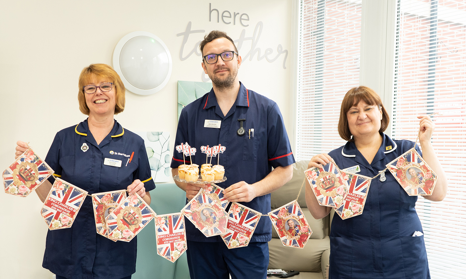 Two women and a man wearing dark blue medical scrubs, holding Coronation-themed bunting in red, blue and white. The man is also holding a tray of cupcakes.
