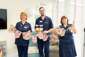 Two women and a man wearing dark blue medical scrubs, holding Coronation-themed bunting in red, blue and white. The man is also holding a tray of cupcakes.