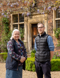 A woman with silver hair and floral dress standing with man in black vest and blue top standing in front of manor house with wisteria.