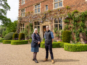 A woman and a man standing in front of a manor house with wisteria, winding shrubbery and plants.