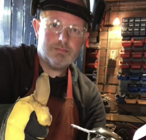 Brett Barker, one of the Artists for the St Barnabas HeART Trail. Man wearing safety goggles and gloves giving thumbs up inside a workshop environment.