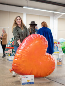 Young brunette woman with orange heart-shaped sculpture, designed to look like a balloon. Part of St Barnabas Hospice's HeART Trail.