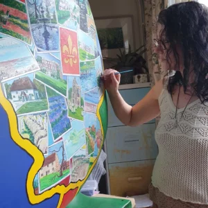 Jem Dunn, one of the Artists for the St Barnabas HeART Trail. Woman with dark hair wearing white crochet top painting colourful heart shaped sculpture.
