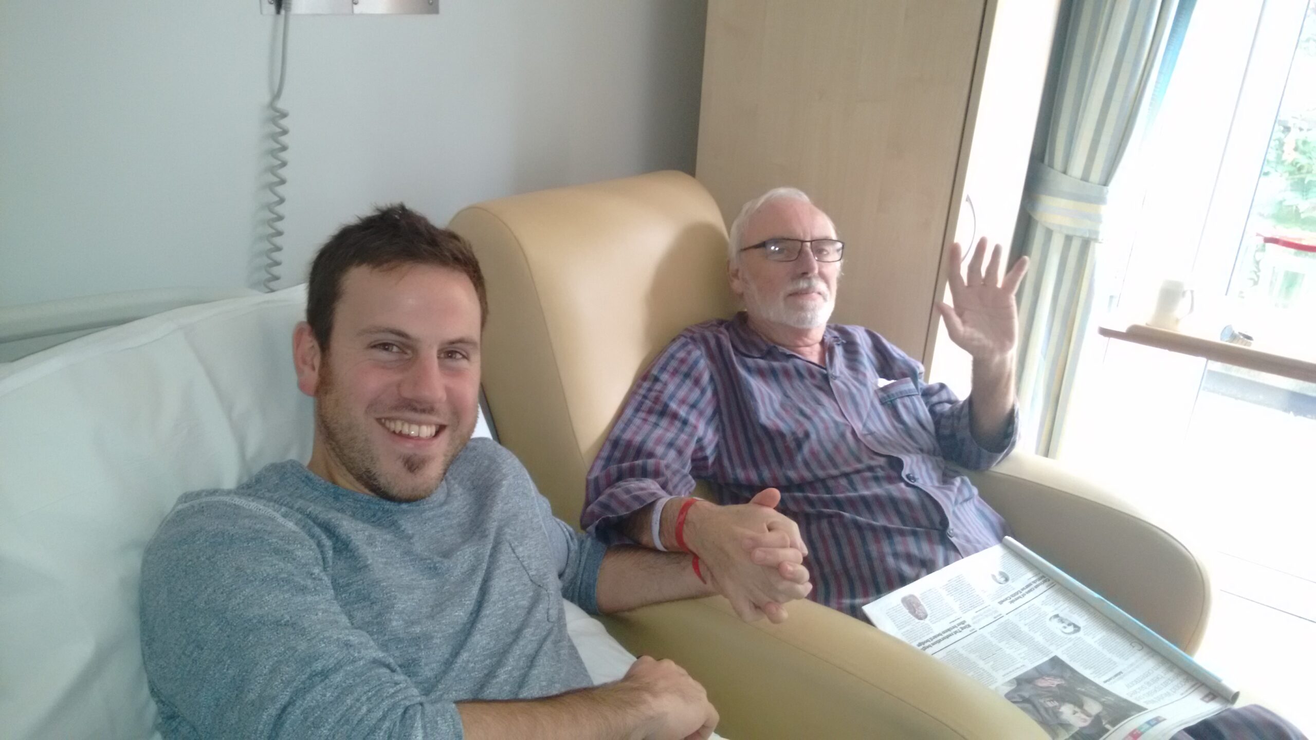A patient at the St Barnabas Inpatient Unit, Steve Hardy,with his son, Jim Hardy. Steve is sat in a recliner bed, with Jim by his side, holding hands.