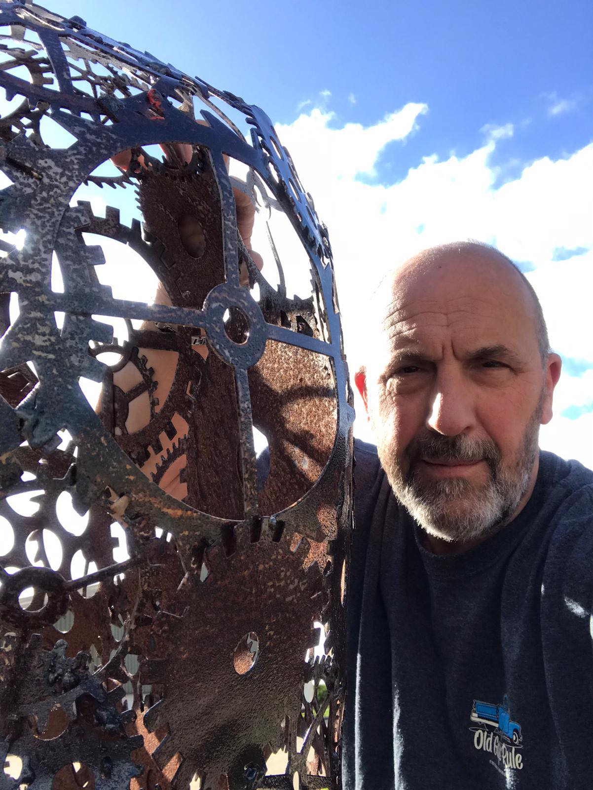Kevin Baumber, one of the Artists for the St Barnabas HeART Trail. Man with beard posing with sculpture depicting industrial cogs made of metal.