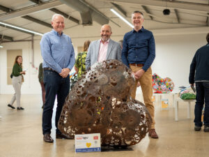 Three men standing with heart-shaped metal sculpture, part of St Barnabas Hospice's HeART Trail..