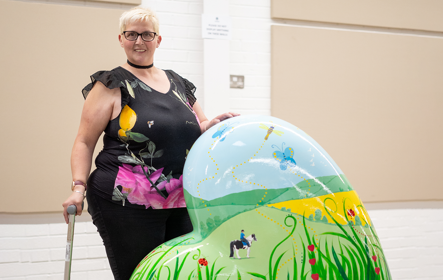 Woman with short blonde hair wearing black top and trousers holding cane. Standing next to colourful heart sculpture depicting horse and rider in field with flowers.