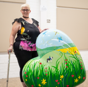 Woman with short blonde hair wearing black top and trousers holding cane. Standing next to colourful heart sculpture depicting horse and rider in field with flowers.
