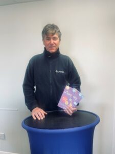Man with silver hair and black fleece jacket and St Barnabas logo, holding purple Lottery leaflets by blue podium