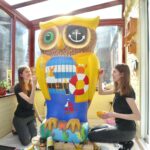 Phillippa and Rachael Corcutt, two of the HeART Trail artists. Two young women with long brown hair, wearing black tops and grey trousers, painting large owl statue to look like a pirate. They are inside a conservatory with brown window frames.