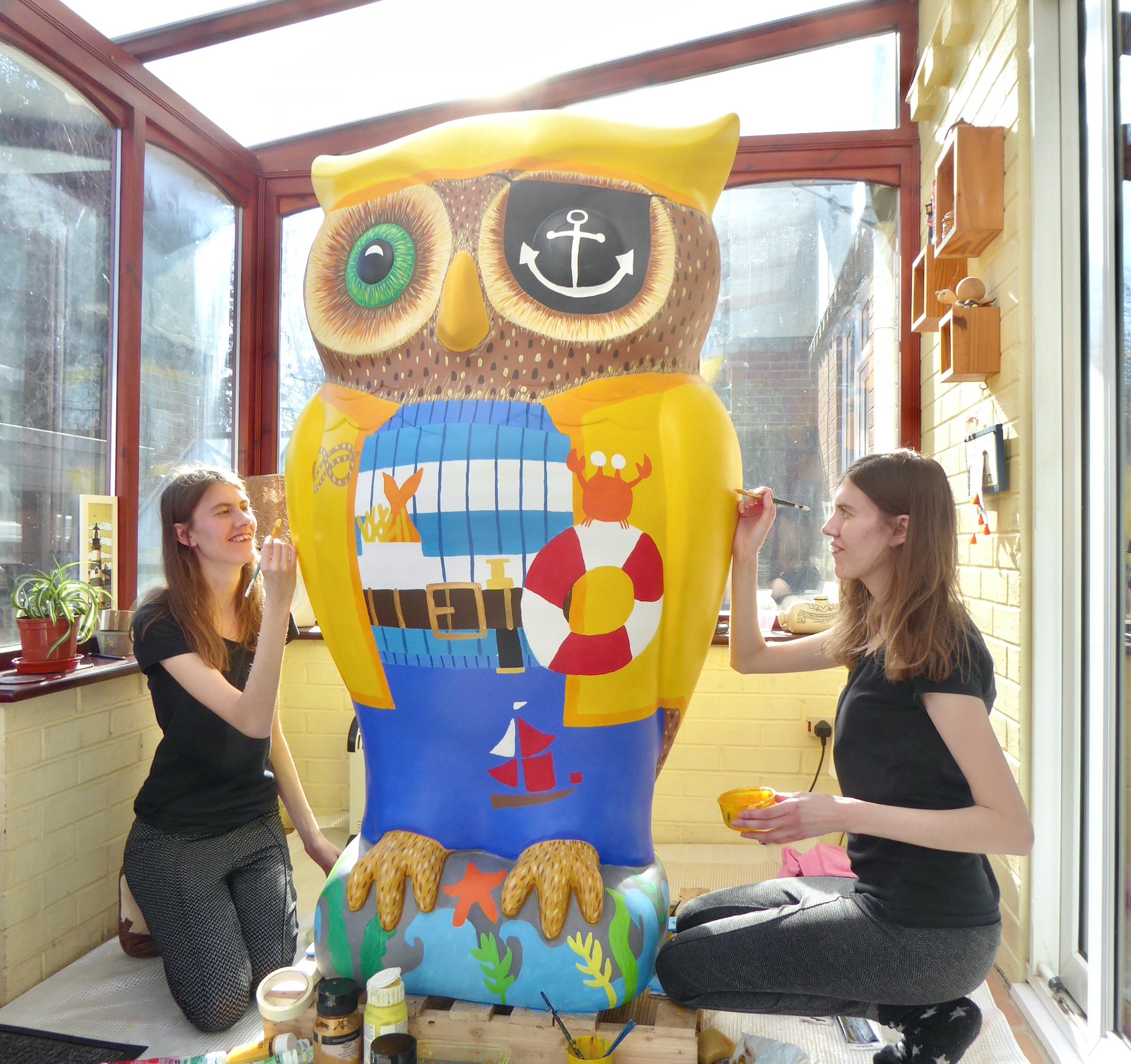 Phillippa and Rachael Corcutt, two of the HeART Trail artists. Two young women with long brown hair, wearing black tops and grey trousers, painting large owl statue to look like a pirate. They are inside a conservatory with brown window frames.