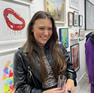 Sian Bristow, one of the Artists for the St Barnabas HeART Trail. Woman with long brown hair wearing leather jacket, smiling with paintings on wall behind her.