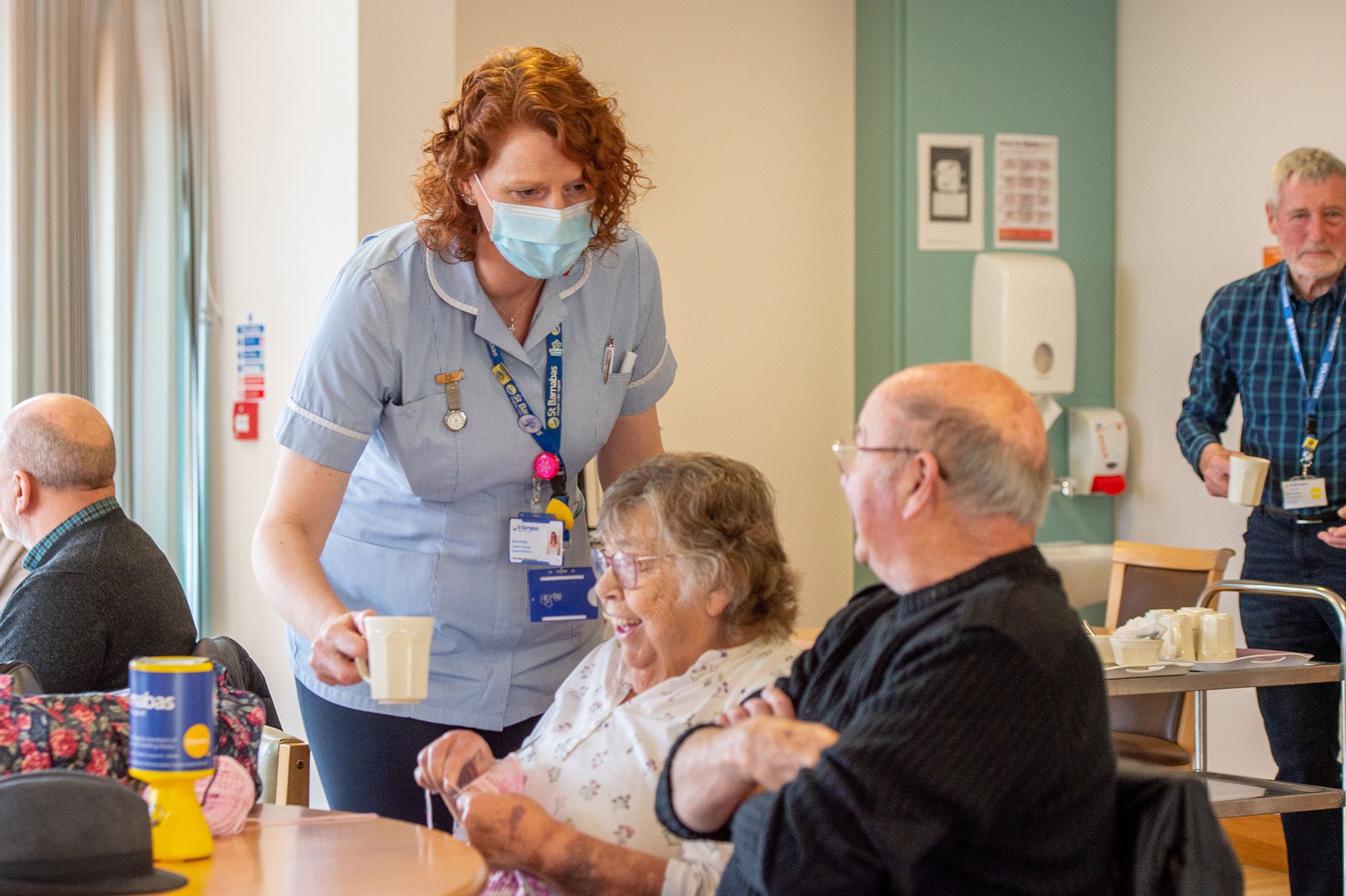 An elderly man and woman, sat at a table, receving a cup of tea from a St Barnabas Health Care Support Worker.