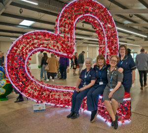 Four women in nurse uniforms sitting on bench of a large red heart-shaped sculpture., part of St Barnabas Hospice's HeART Trail.