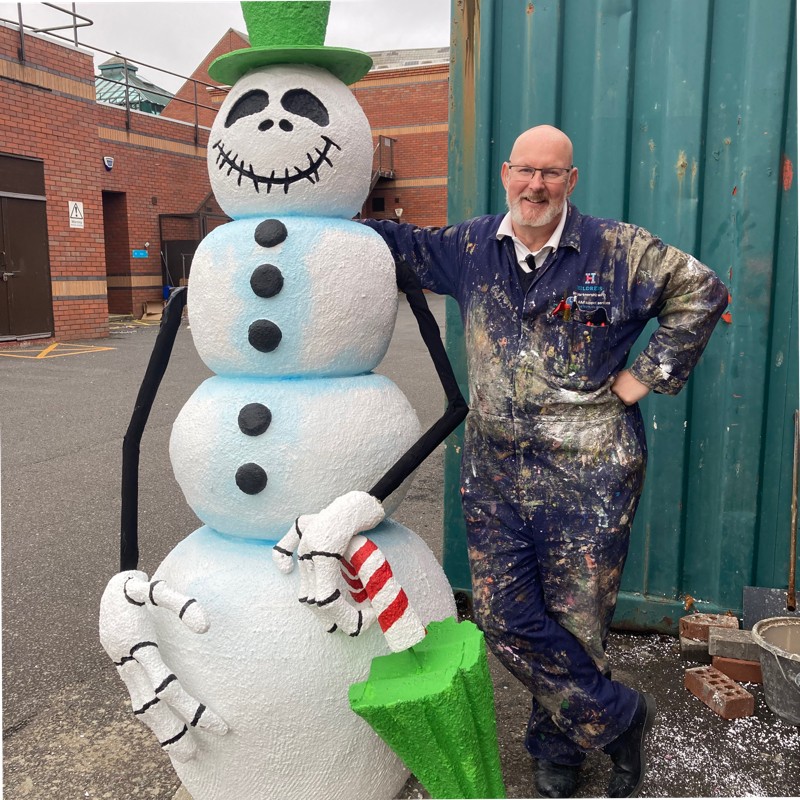Steve Andrews, one of the Artists for the St Barnabas HeART Trail. Man dressed in paint-stained blue coveralls standing by large statue of snowman.