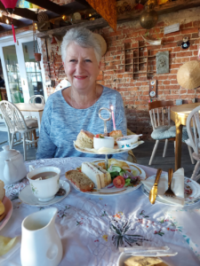 Grey haired woman wearing blue at a table with afternoon tea laid out.