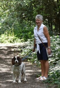 Grey haired woman wearing white and black, with a spaniel dog outside in the woods