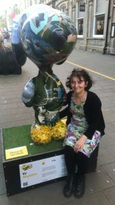 Kathleen Smith, one of the Artists for the St Barnabas HeART Trail. Woman with short black hair, wearing black and tropical print clothing, sitting by a statue painted with wildlife theme.