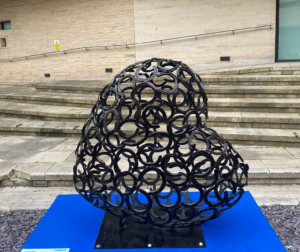 Heart-shaped sculpture made of horseshoes on blue plinth in front of steps of building, part of the St Barnabas HeART Trail