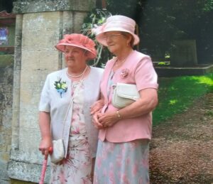 Two elderly women, stood in front of a church yard, at a wedding. Both are dressed in formal wedding attire, in pink and blue colour schemes, with matching hats.