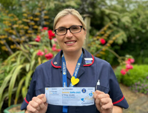 Nurse in dark blue uniform and light blue lanyard, holding a raffle ticket in garden