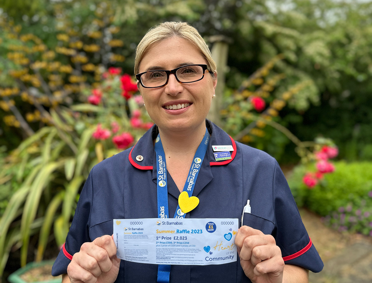 Nurse in dark blue uniform and light blue lanyard, holding a raffle ticket in garden