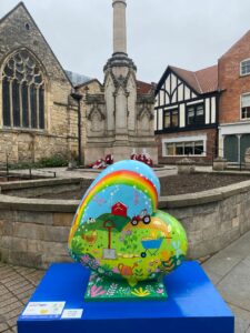 Brightly coloured heart-shaped sculpture with rainbow, tractor and agricultural design on blue plinth in front of St Benedicts Square in Lincoln, part of the St Barnabas HeART Trail