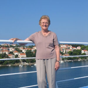 An elderly women, wearing a pink top and beige trousers, standing against a railing of a boat, sailing past a Mediterranean town, in sunny weather.